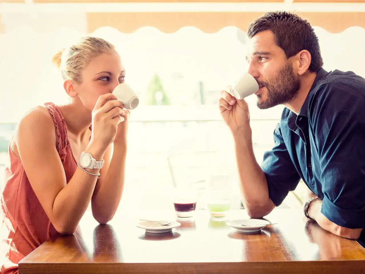 Un uomo innamorato in segreto, con lo sguardo perso, beve un caffè mentre osserva la donna di fronte a lui.