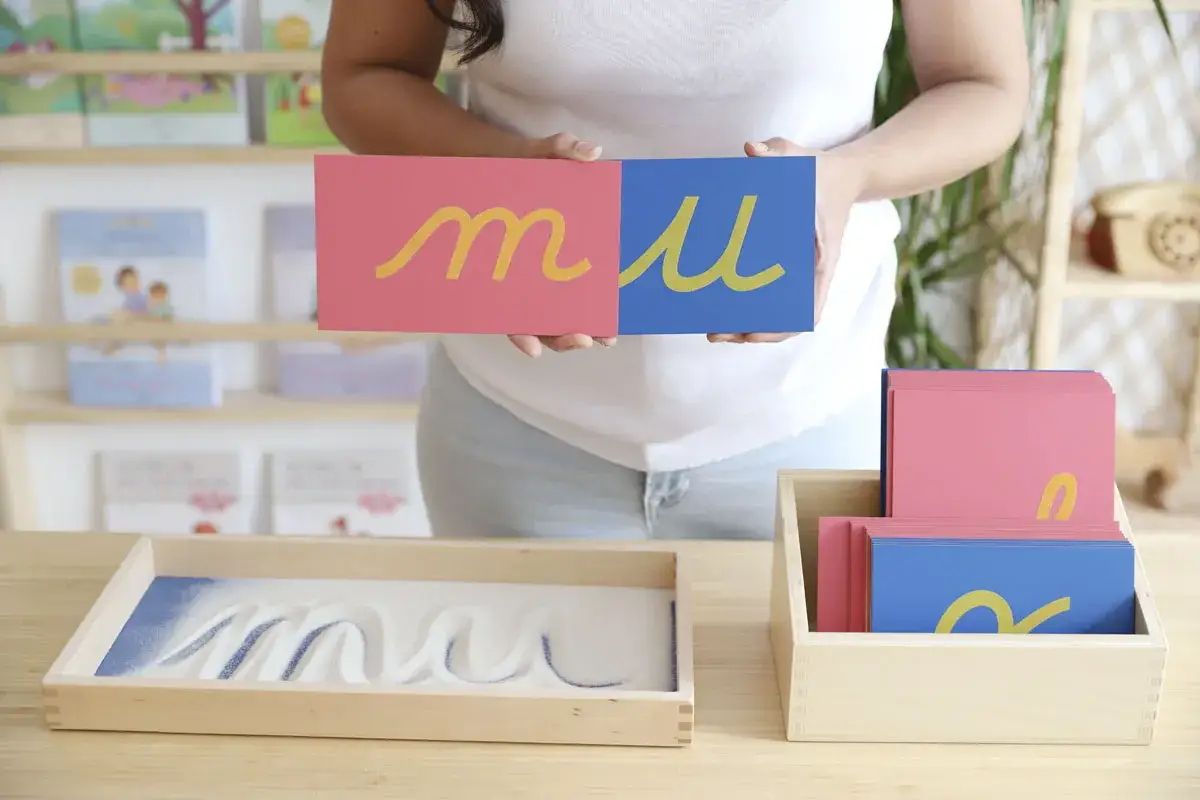 Niño practica preescritura montessori trazando letras en arena. Tarjetas con letras doradas sobre fondo rosa y azul.