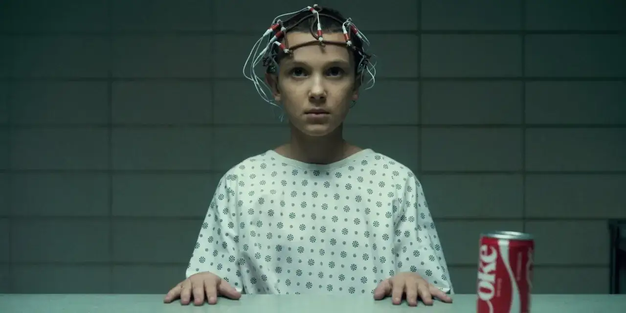 Nastka z Stranger Things, z elektrodami na głowie, w szpitalnej koszuli, obok puszki Coca-Coli.