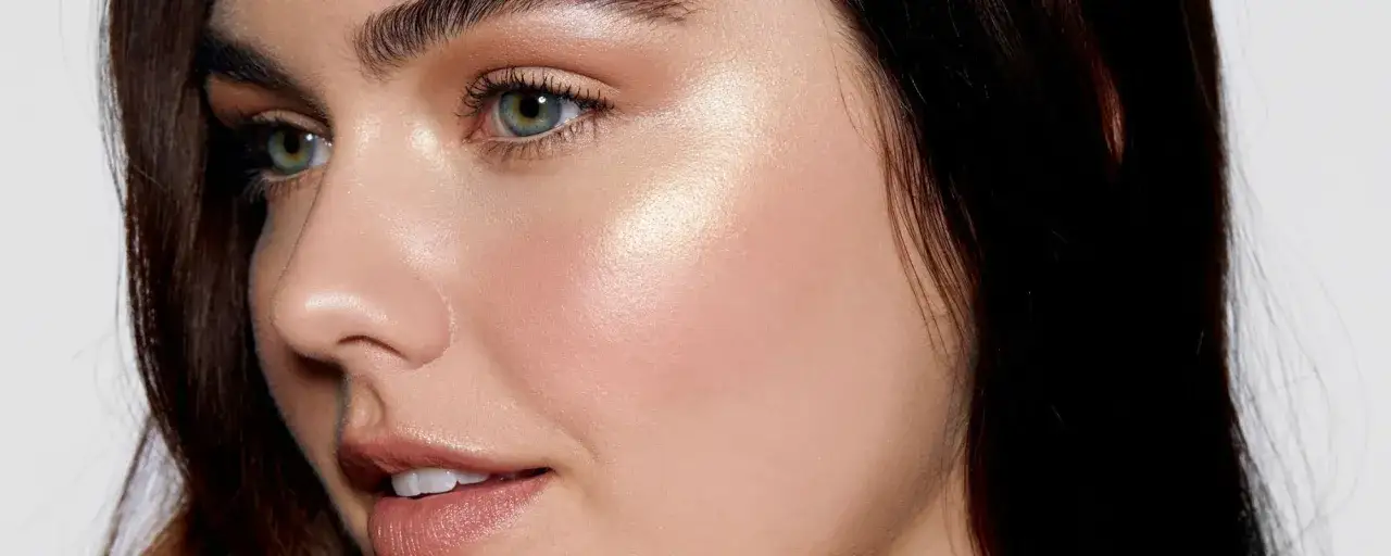 En kvinna med gröna ögon och mörkt hår som har fått en glödande finish genom att applicera highlighter på kindbenen.