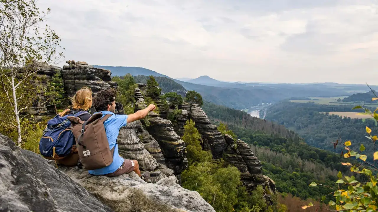Zwei Wanderer genie&szlig;en die atemberaubende Deutschland Landschaft von einem Felsvorsprung aus. Der Mann zeigt in die Ferne, wo sich ein Fluss durch gr&uuml;ne T&auml;ler schl&auml;ngelt.