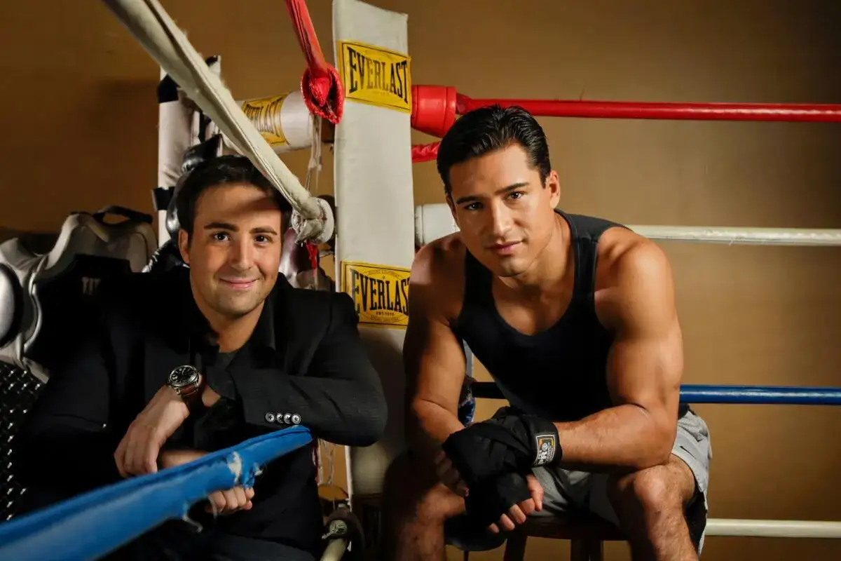 Mario Lopez in boxing gear, ready for a match, sits beside a man in a suit in a boxing ring.