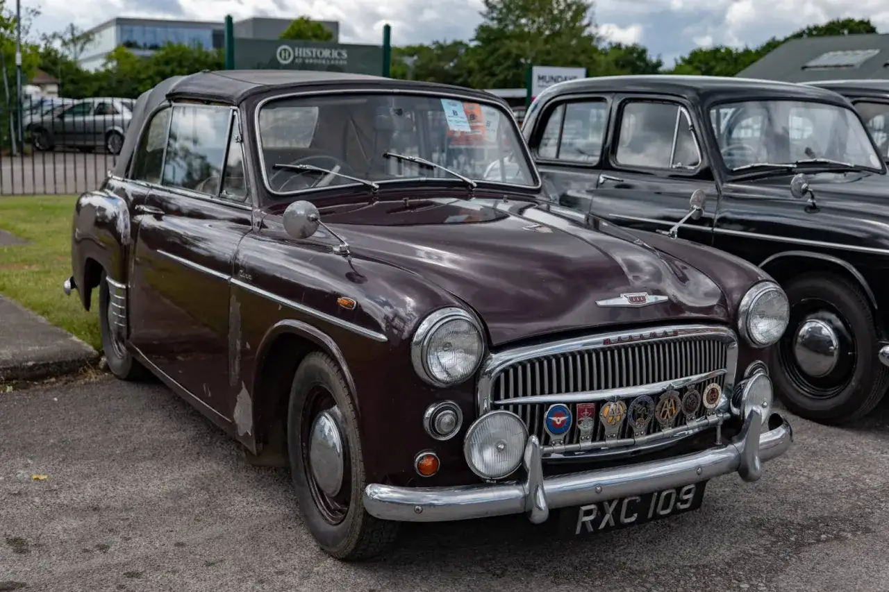 En mörklila Hillman-bil med cabriolettak står parkerad bredvid en svart bil. Bilen har en kromad grill med flera märken.