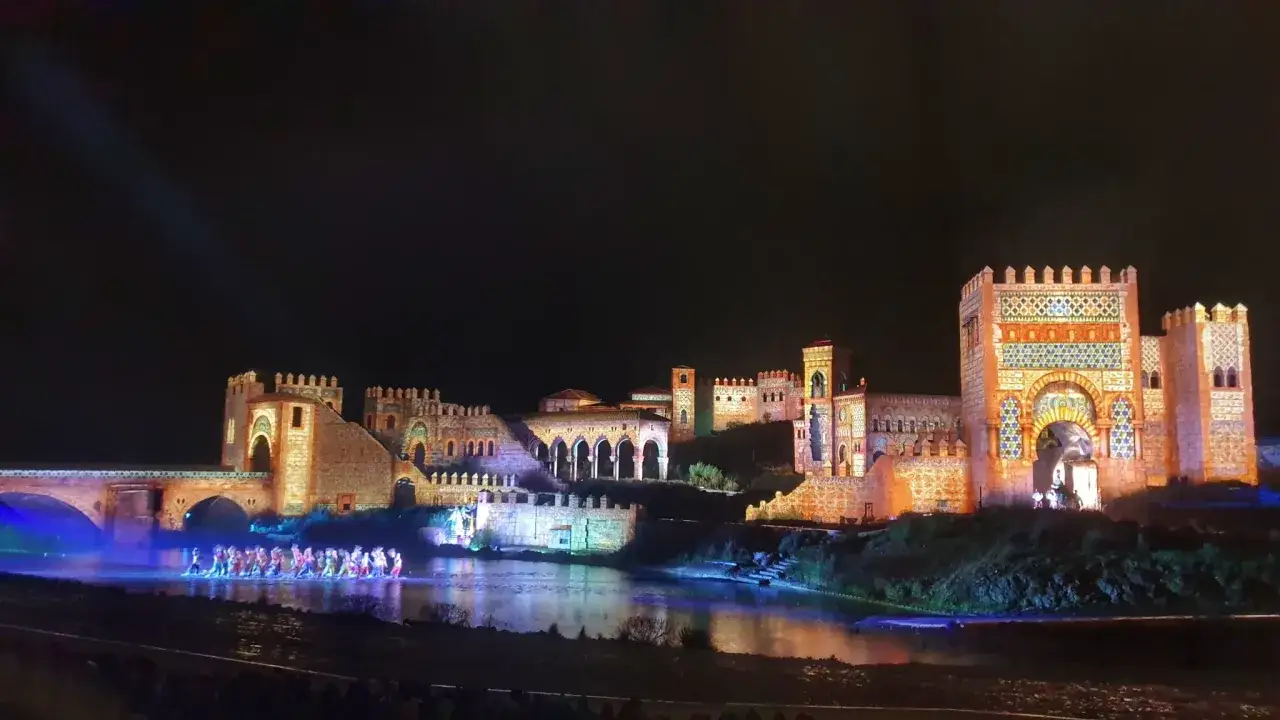 Espect&aacute;culo nocturno en Puy du Fou Francia: un puente y castillos iluminados con bailarines en el agua.