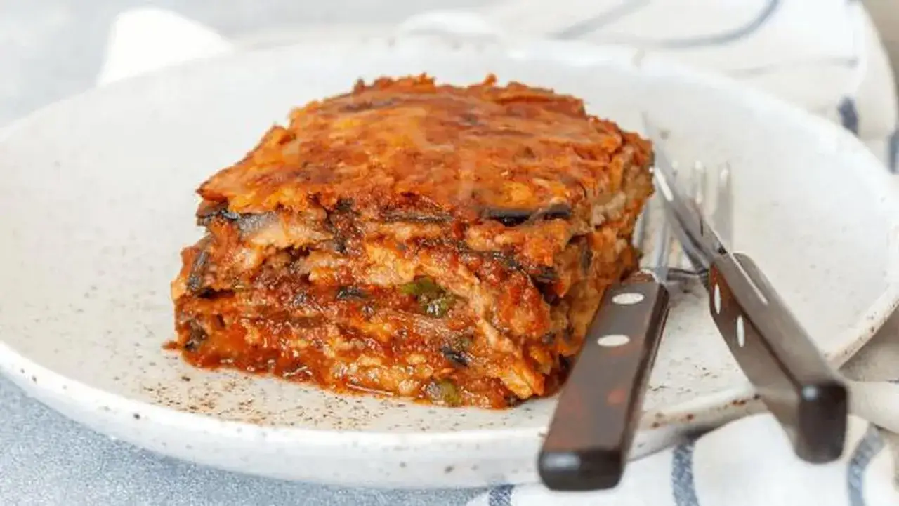 Porci&oacute;n de pastel de berenjenas en capas, con salsa de tomate y queso, servida en un plato blanco con cubiertos.