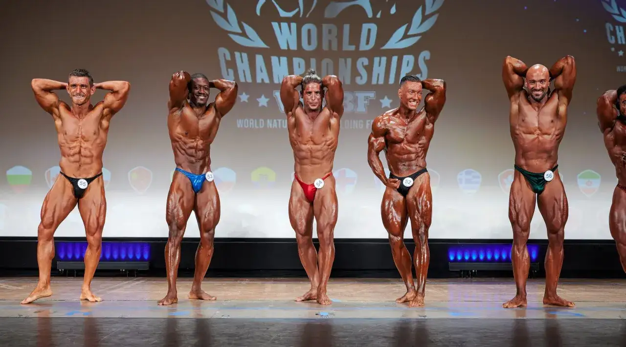 F&uuml;nf muskul&ouml;se M&auml;nner pr&auml;sentieren ihre K&ouml;rper bei einem Bodybuilding Wettkampf. Sie stehen auf einer B&uuml;hne vor einem Banner mit der Aufschrift 