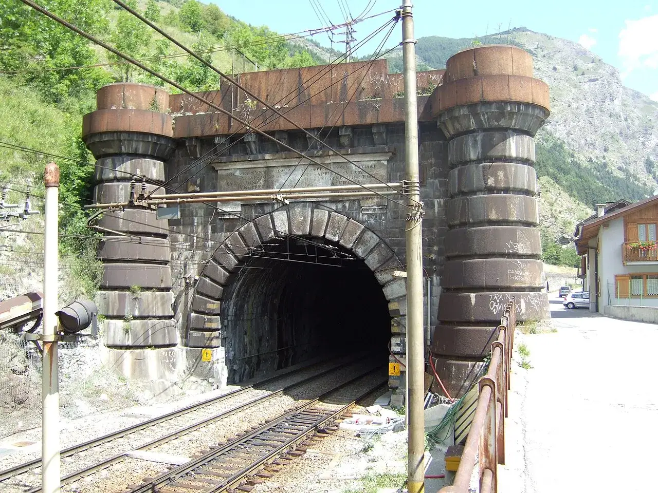 Kamienne filary i łuk wejściowy do tunelu Frejus, z widocznymi torami kolejowymi i górami w tle.