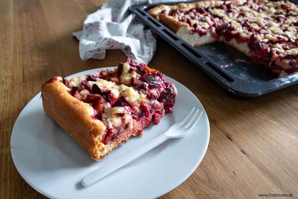 Ein St&uuml;ck Zwetschgendatschi mit Streuseln auf einem Teller, daneben der ganze Kuchen im Blech. Ein leckeres Zwetschgendatschi Rezept zum Nachbacken.