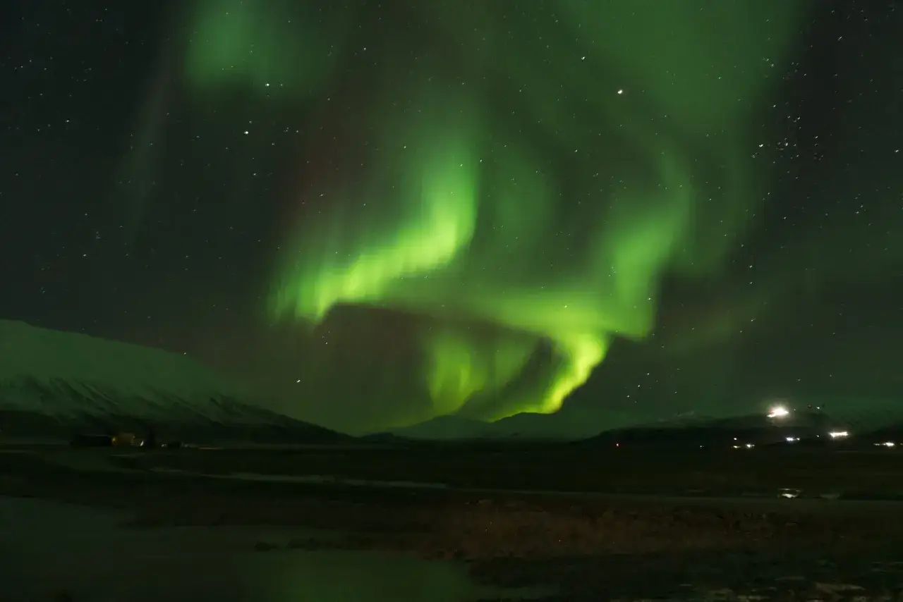 Zielone zorze polarne tańczą na nocnym niebie nad ośnieżonymi górami i niewielką osadą.