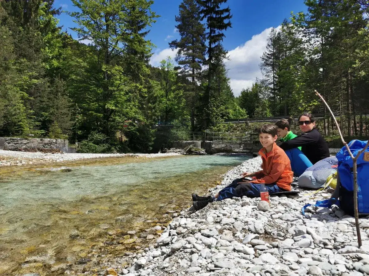 Familie genießt Urlaub in Bayern am Fluss. Kinder und Mutter sitzen am Kiesstrand, umgeben von grünen Wäldern.