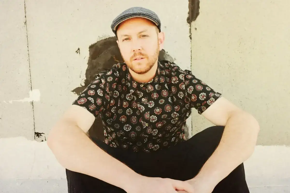 Matt Simons, nach dem Erdrutsch, sitzt l&auml;ssig in einem gemusterten Hemd und Schieberm&uuml;tze vor einer rauen Wand.