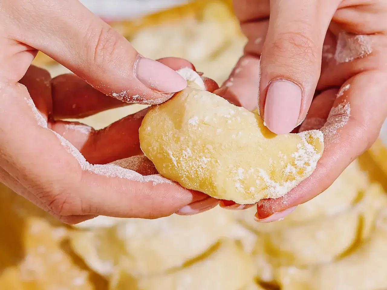 Dłonie z różowymi paznokciami formują ciasto na pierogi, posypane mąką. W tle widać gotowe pierogi.
