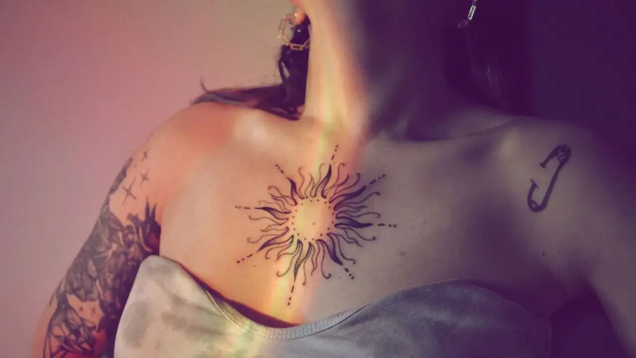 Ein strahlendes Seelensymbol-Tattoo auf der Brust, umgeben von zarten Punkten und Strahlen. Ein kleiner Sicherheitsnadel-Tattoo auf der Schulter.