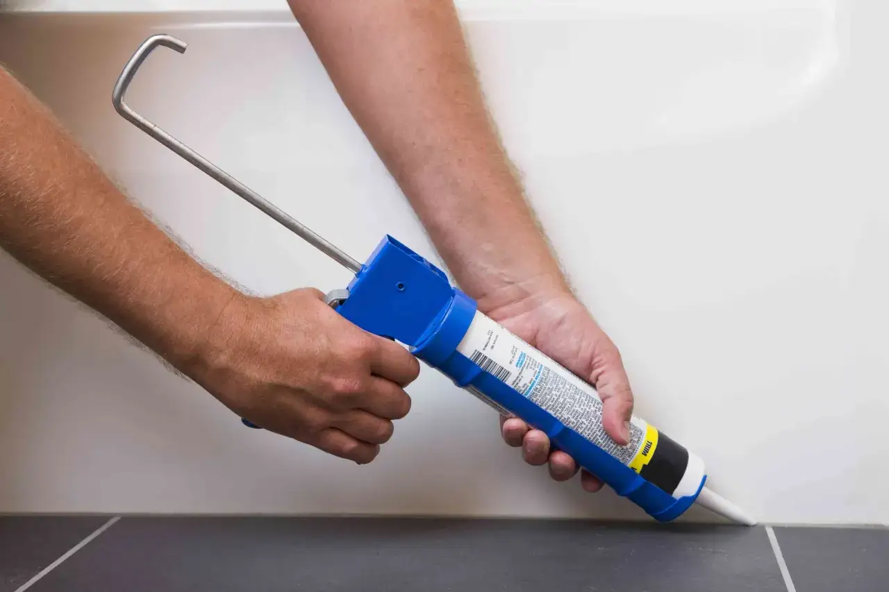 Hands using a caulk gun to apply sealant along the edge of a bathtub, demonstrating how to caulk a bathtub for a watertight seal.
