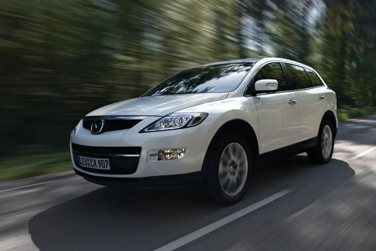 Biała Mazda CX-9 pędzi leśną drogą, rozmazując zieleń drzew.