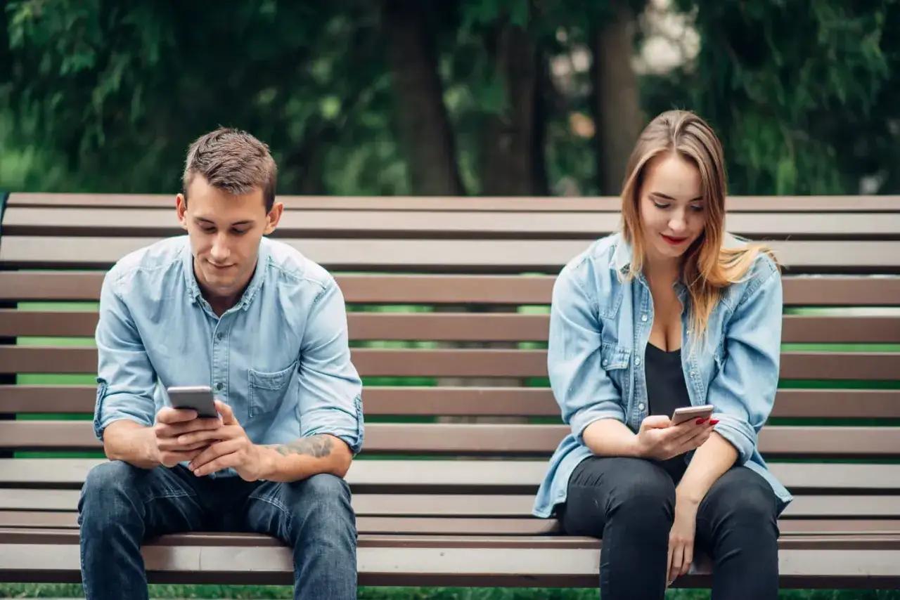 Un couple assis sur un banc, chacun absorbé par son téléphone. Un exemple de l'effet boomerang en amour, où la distance numérique crée une distance réelle.