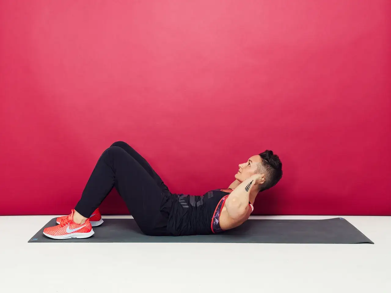 Kobieta wykonuje ćwiczenia na brzuch, znane jako **situps**. Leży na macie, z nogami ugiętymi w kolanach, ręce splecione za głową.