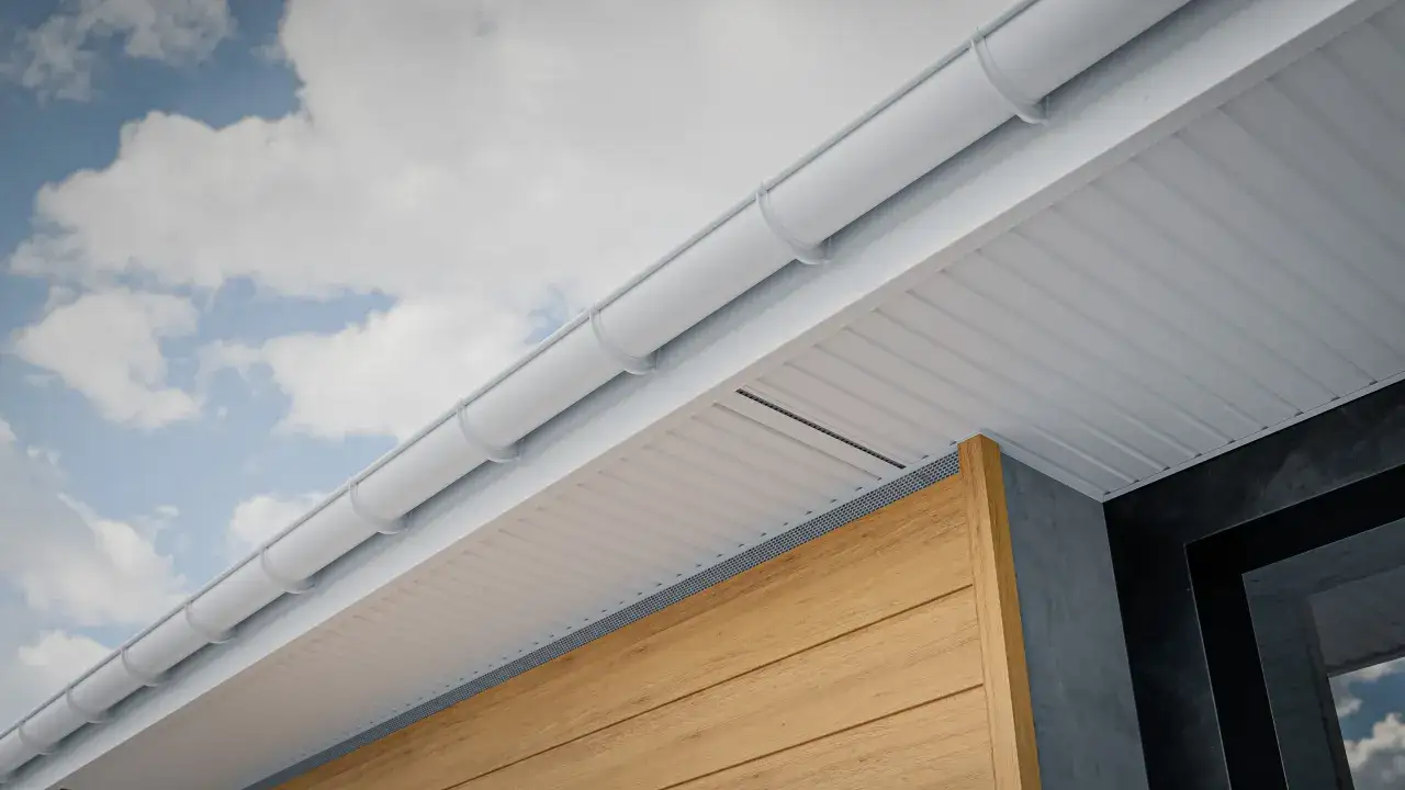 Gouttière blanche et soffite blanc rayé, sous un ciel bleu nuageux. La toiture froide est bien visible, avec un bardage en bois clair et un mur sombre.