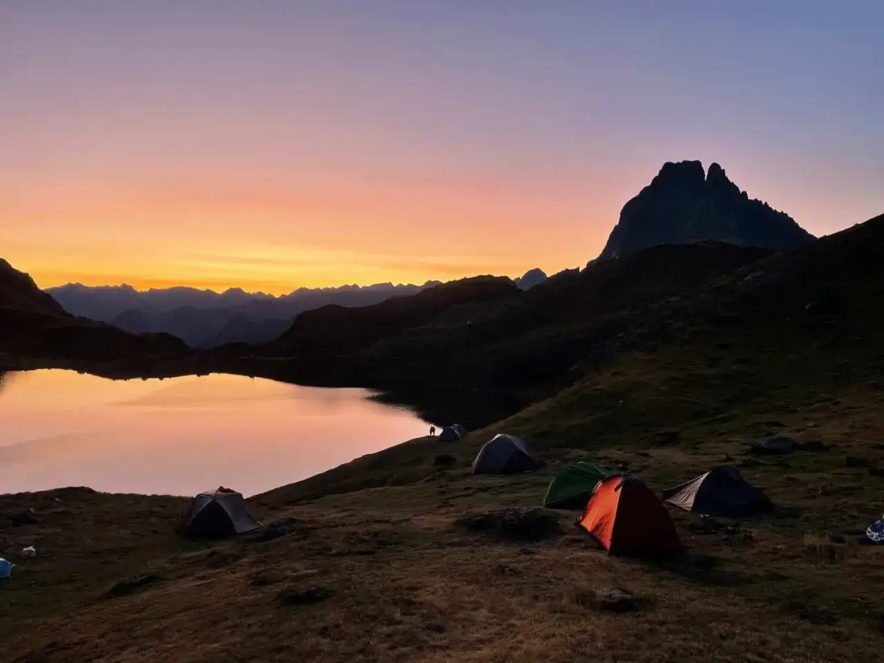 Un bivouac paisible au bord d'un lac de montagne, sous un ciel aux couleurs chaudes du crépuscule. Plusieurs tentes sont installées sur la pente herbeuse.