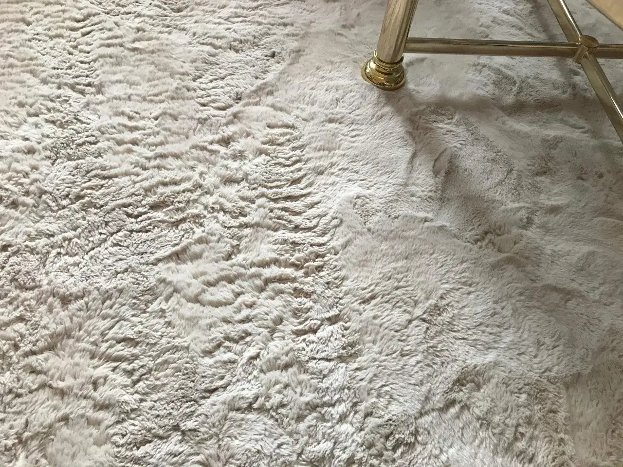 Ein heller, verfilzter Kunstfell Teppich mit einem goldenen Möbelfuß.