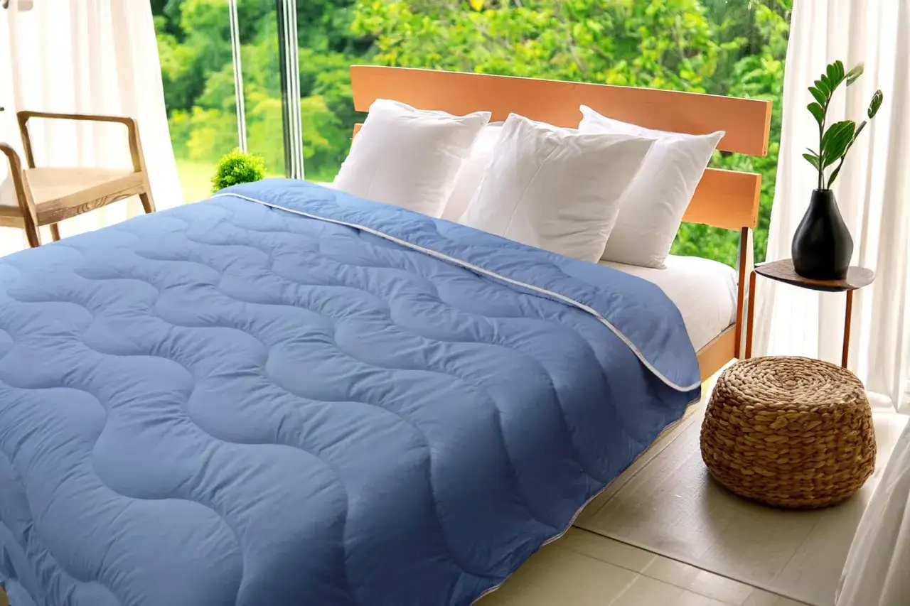 Une couette bleue l&eacute;g&egrave;re, parfaite pour l'&eacute;t&eacute;, est &eacute;tal&eacute;e sur un lit. La fen&ecirc;tre laisse voir une v&eacute;g&eacute;tation luxuriante.