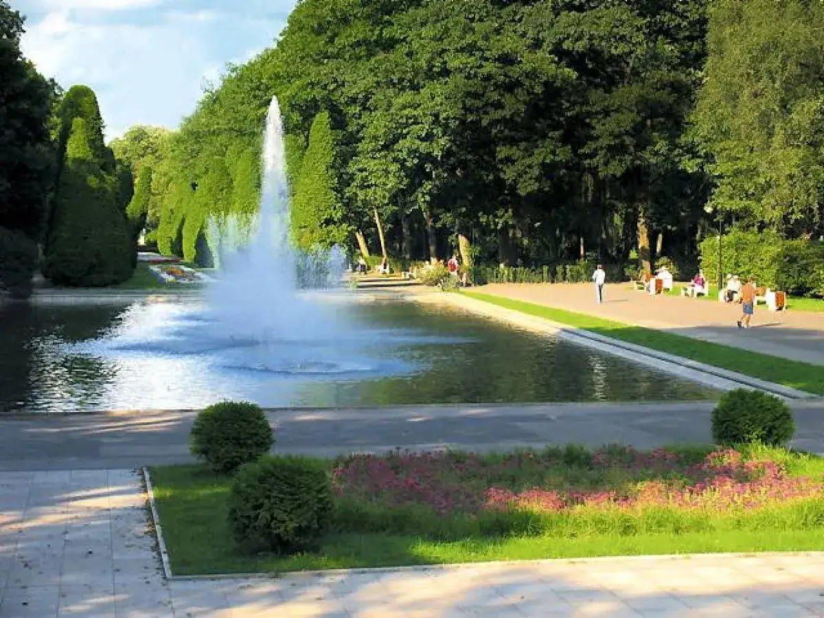 Parkowa fontanna w stylu versailles, otoczona zielenią i alejkami, gdzie spacerują ludzie.