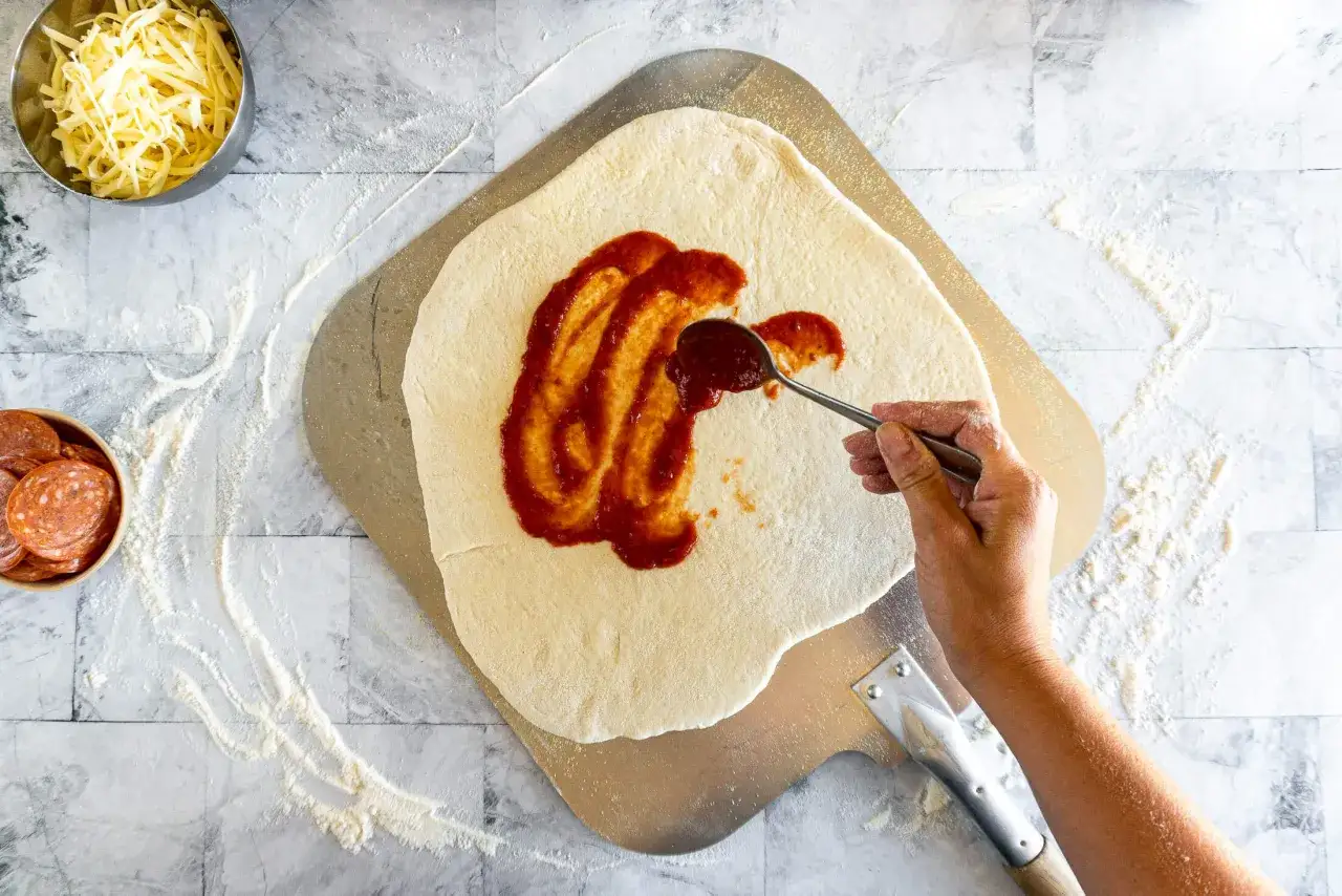 Schneller Pizzateig mit Quark wird mit Tomatensauce bestrichen. Daneben liegen geriebener Käse und Salamischeiben.