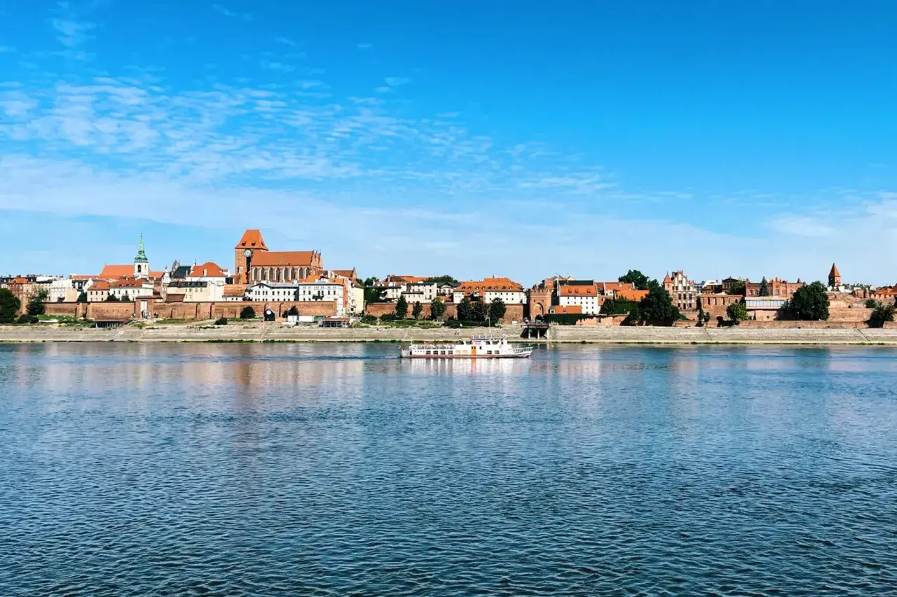Widok na zabytkowe Toruń atrakcje z Wisły. Statek wycieczkowy płynie po rzece, a w tle widać katedrę i kamienice.