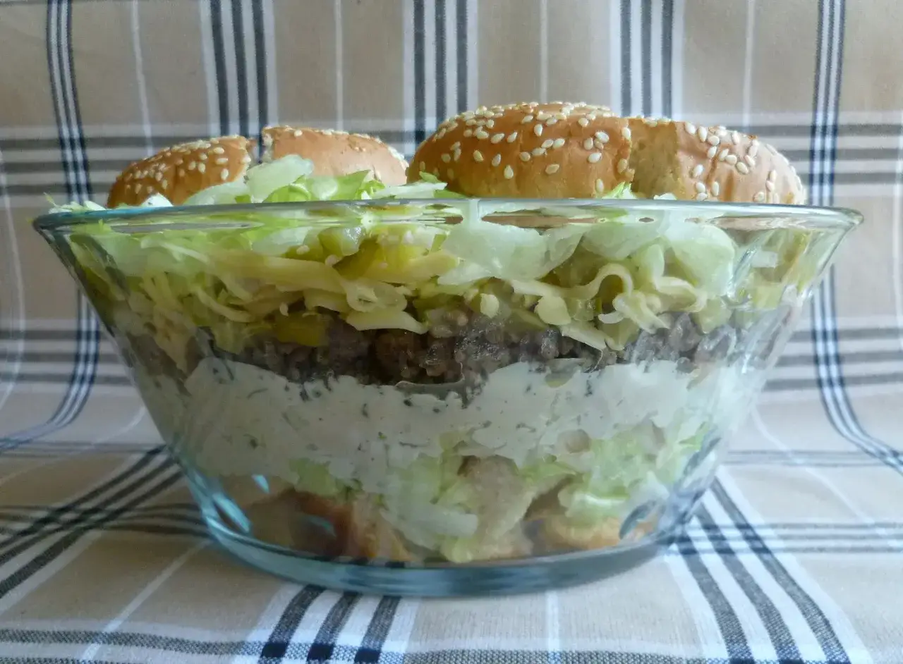 Warstwowa sałatka big mac w szklanej misie, z bułkami na wierzchu.