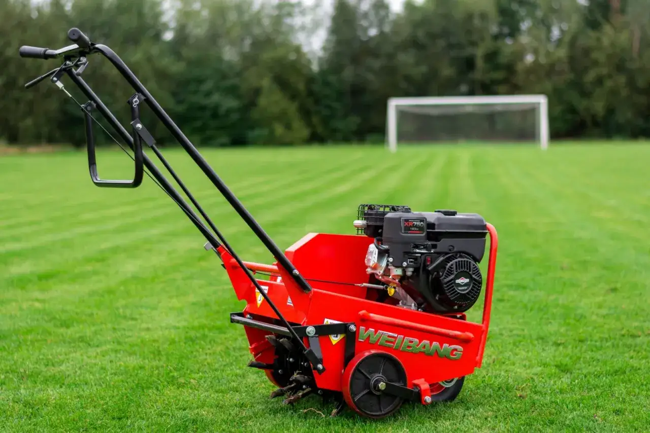 Czerwony aerator do glebogryzarki Weidemann z silnikiem XR750 na zielonej trawie, w tle boisko piłkarskie.