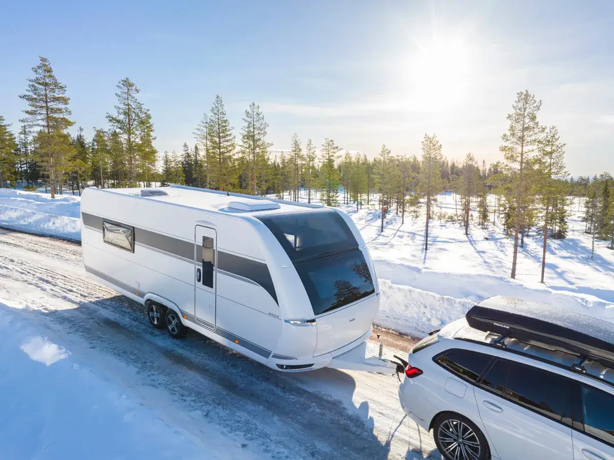Vit husvagn dras av en bil på en snöig väg i skogen. Frågan är om fyrhjulsdrift eller inte är nödvändigt för denna resa.