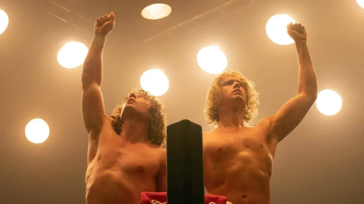 Zwei muskulöse Männer mit langen, blonden Haaren heben triumphierend die Fäuste unter Scheinwerfern. Sie sind Teil der Wrestling-Brüder, die in