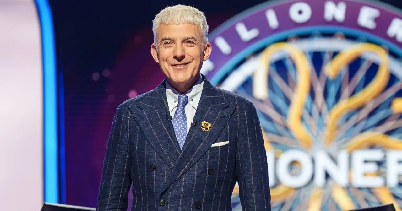 Uśmiechnięty Hubert Urbański w garniturze, gotowy do rozpoczęcia programu Milionerzy.