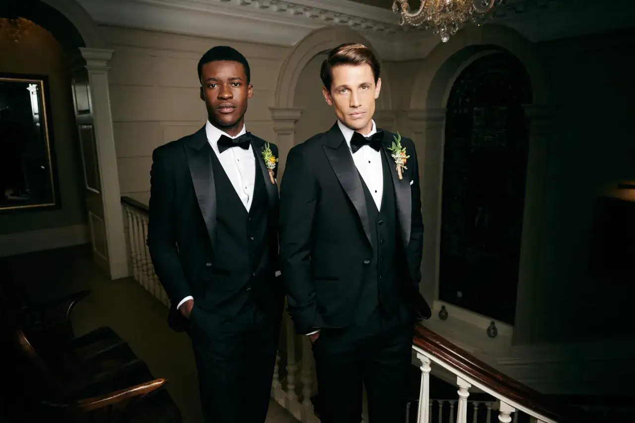 Two men in sharp tuxedos, embodying the gala dinner dress code male. They stand on a grand staircase, ready for an elegant evening.