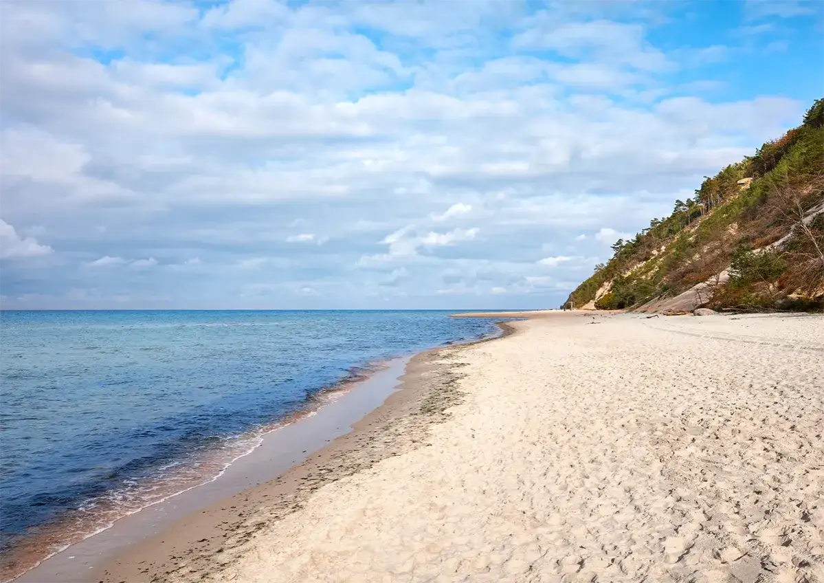 Plaża w Międzyzdrojach z widokiem na zalesione zbocze, zwaną Kawczą Górą. Spokojne morze i piaszczysty brzeg.