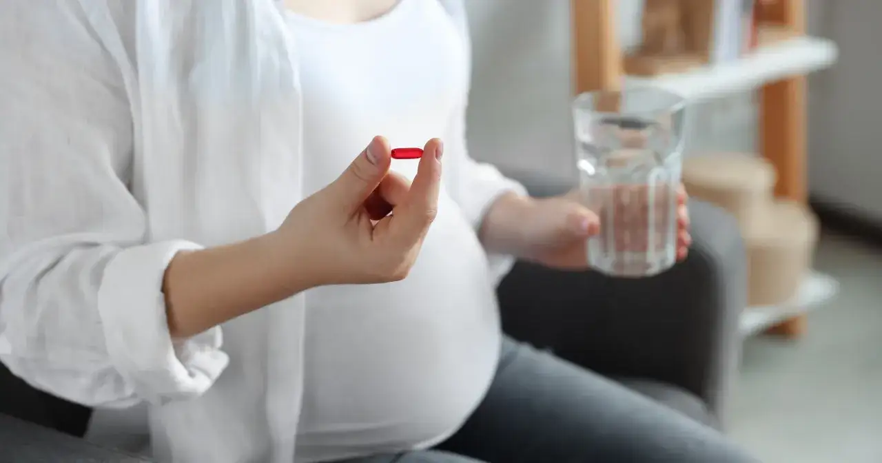 Kobieta w białej koszuli, w 6 tygodniu ciąży, trzyma czerwoną kapsułkę i szklankę wody.