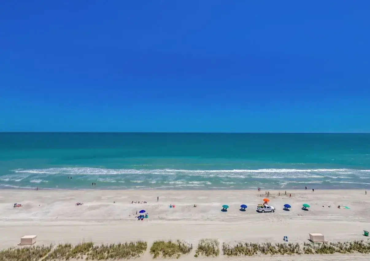 Piękny, **ocean blue** w połączeniu z białym piaskiem i błękitem nieba. Ludzie odpoczywają na plaży, niektórzy kąpią się w morzu.