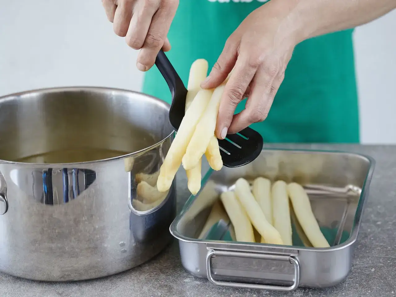 Hände nehmen gekochten Spargel aus einem Topf und legen ihn in eine Auflaufform. Perfekt für Pfannkuchen mit Spargel!