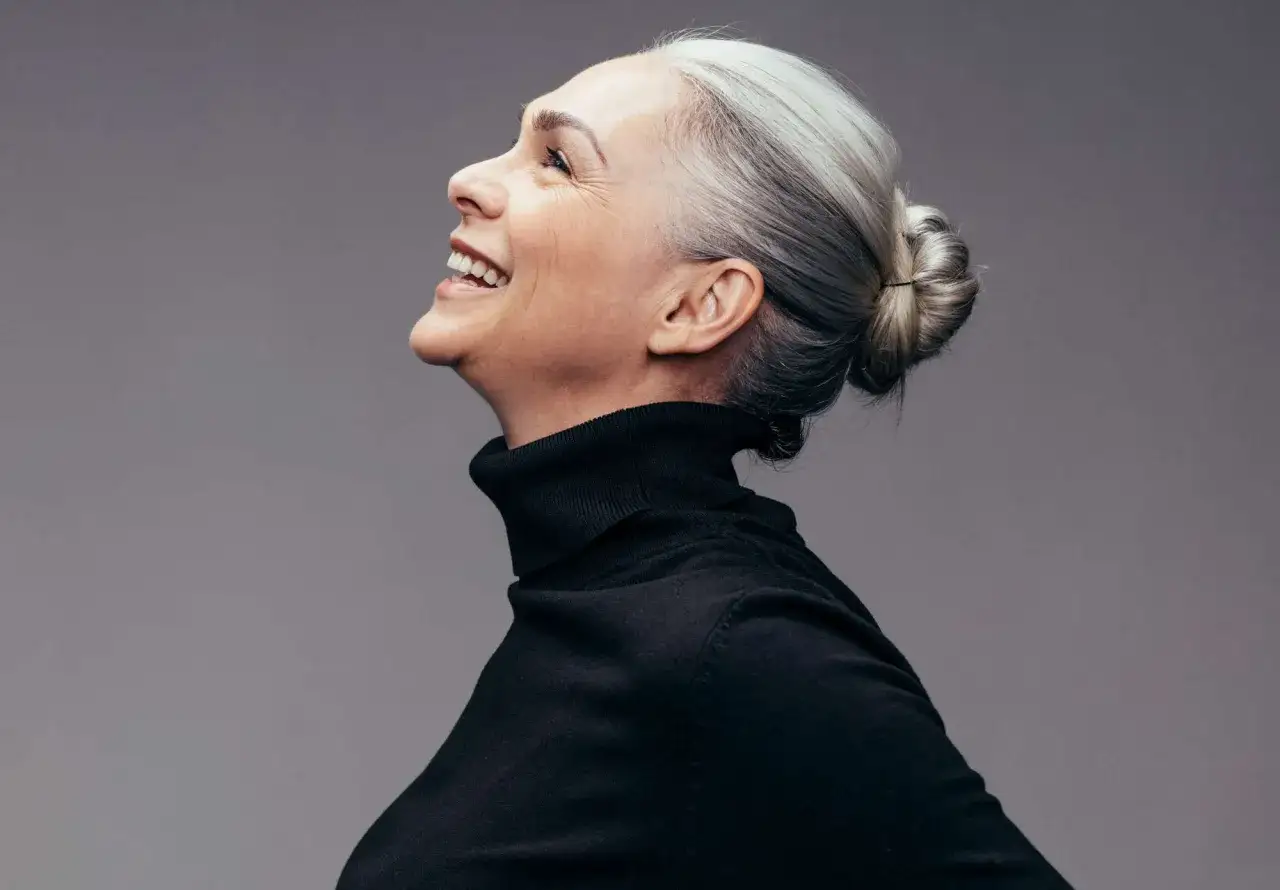 Donna sorridente con capelli bianchi raccolti in uno chignon, indossa un dolcevita nero. Un esempio di come vestirsi con i capelli bianchi con stile.
