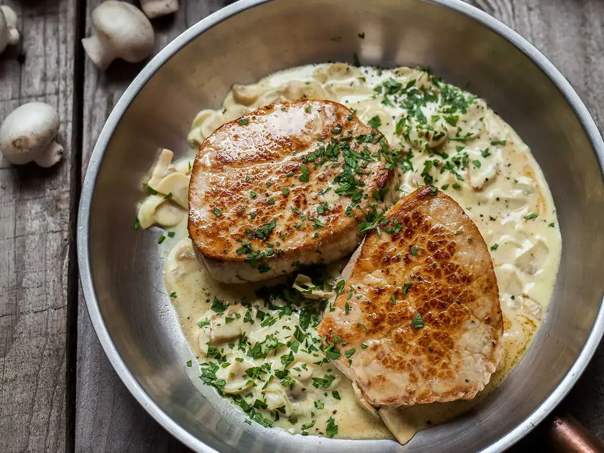 Deux grenadins de veau poêlés, nappés d'une sauce crémeuse aux champignons et parsemés de persil frais, dans une poêle en métal.