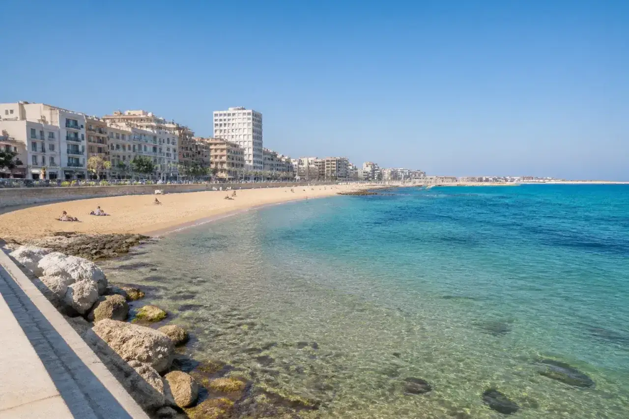 Ein sonniger bari strand mit klarem, türkisfarbenem Wasser und einer Reihe von Gebäuden im Hintergrund.