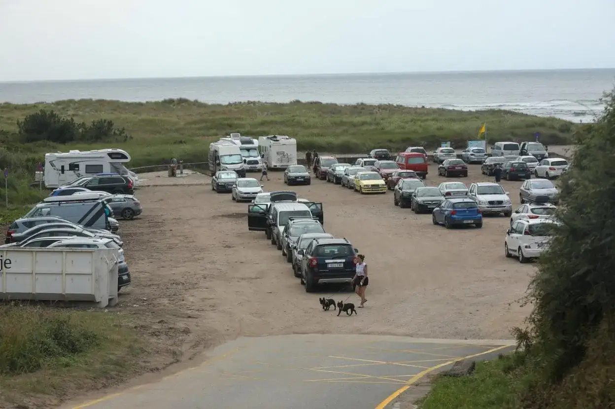 Estacionamiento lleno cerca del playón de bayas. Coches y autocaravanas aparcados en la arena, con la playa y el mar al fondo.