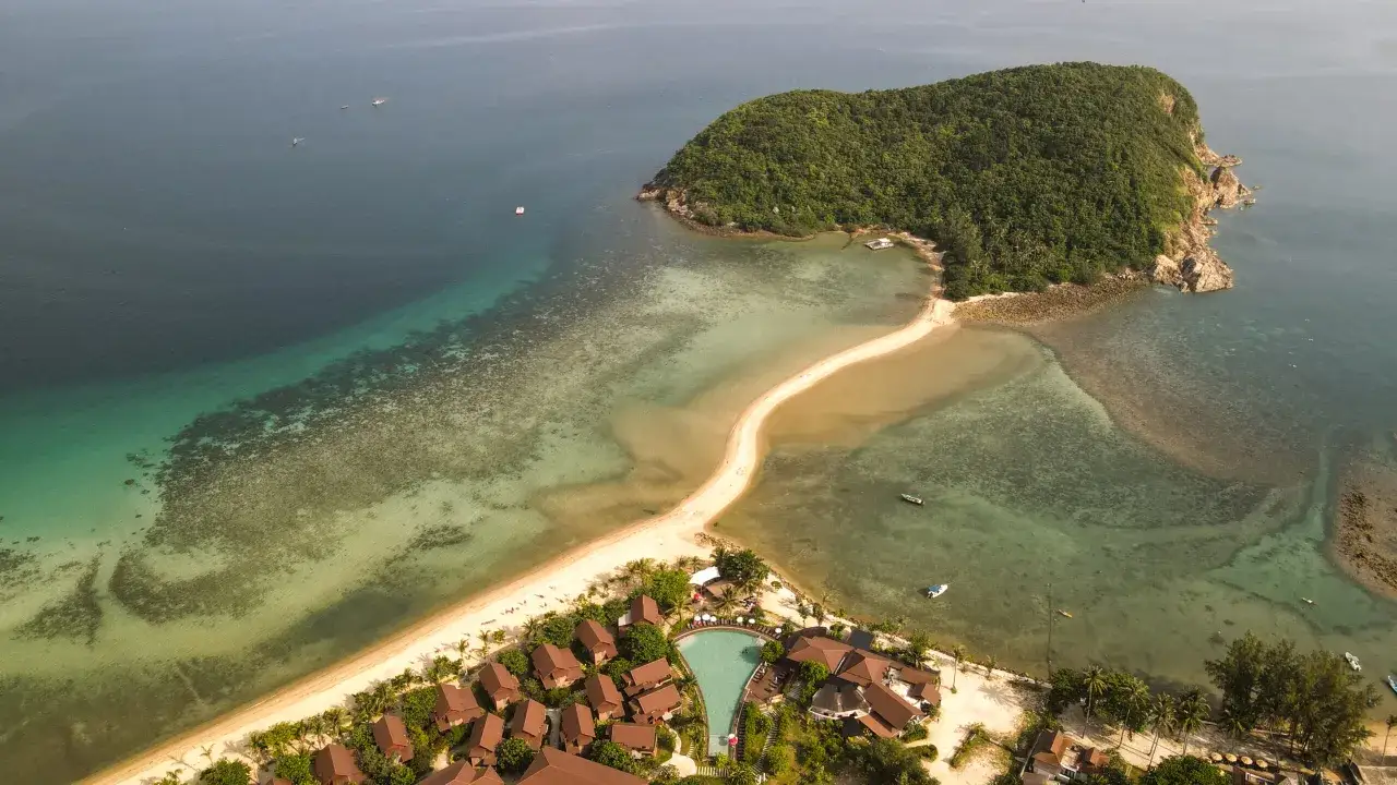 En sandbank leder till en grönskande ö vid Koh Phangan, Thailand. En resort med pool och bungalows ligger vid stranden.