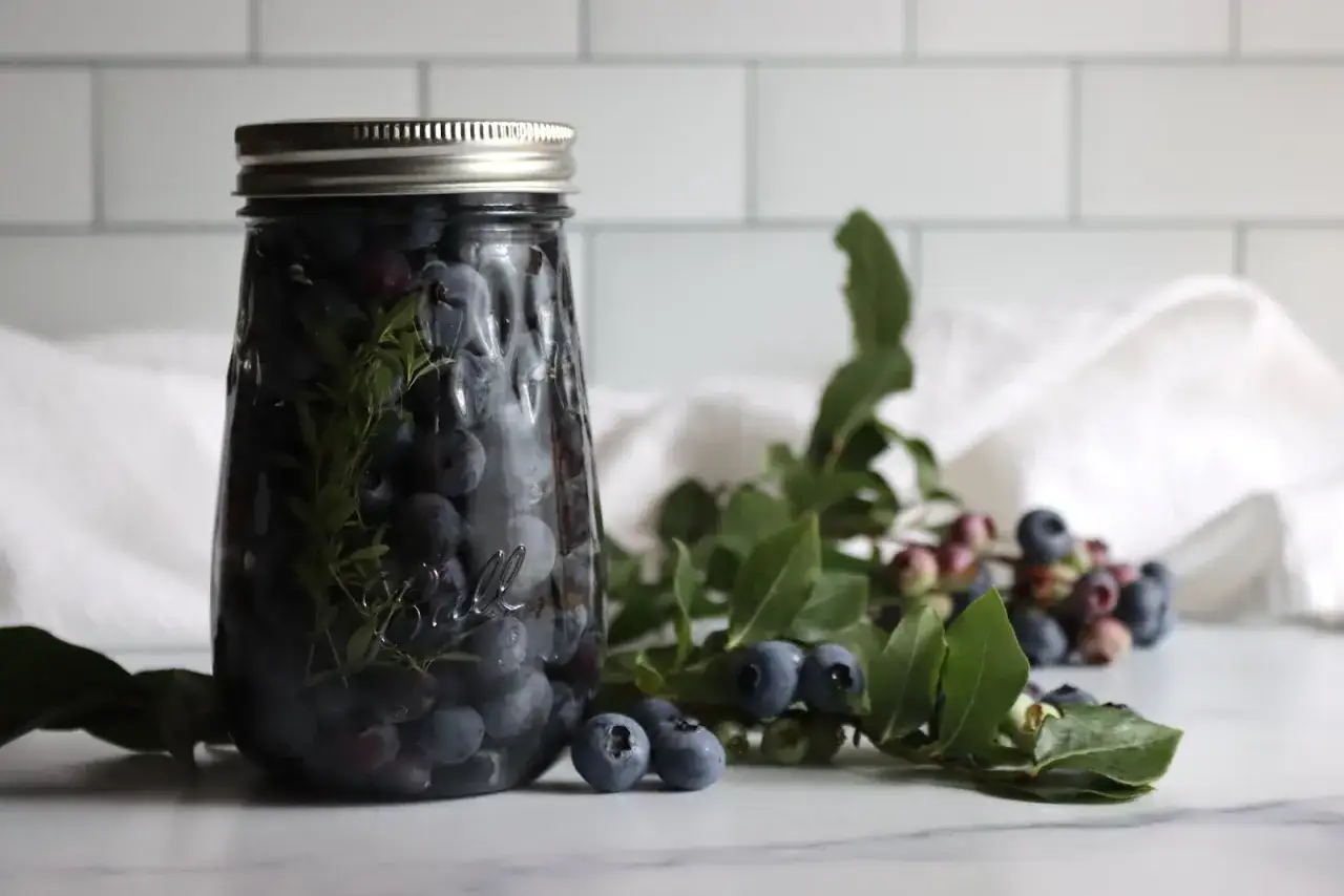 Ein Glas wird mit Heidelbeeren und Kräutern gefüllt. Frische Beeren liegen daneben.
