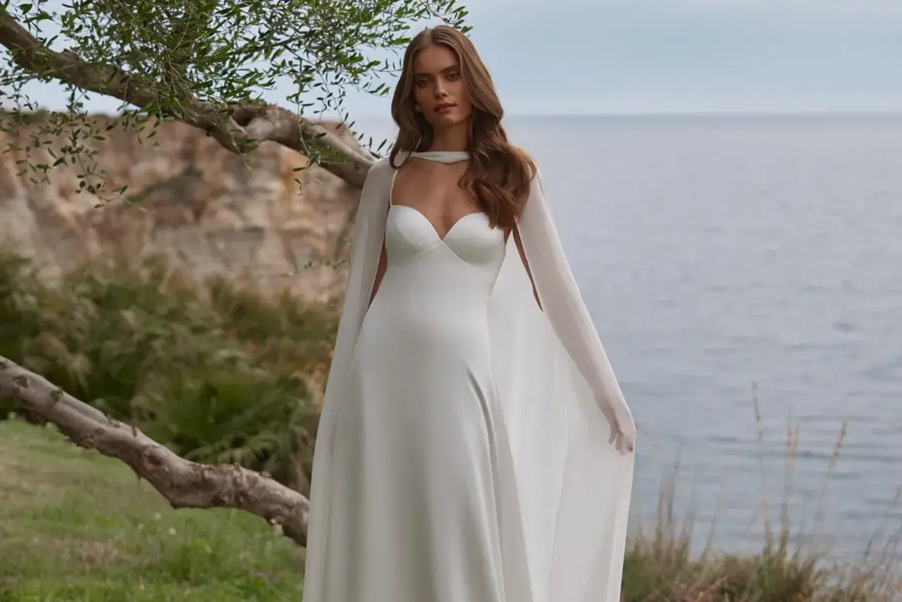 Elegancka panna młoda w minimalistycznej sukni ślubnej z peleryną, pozująca nad morzem.