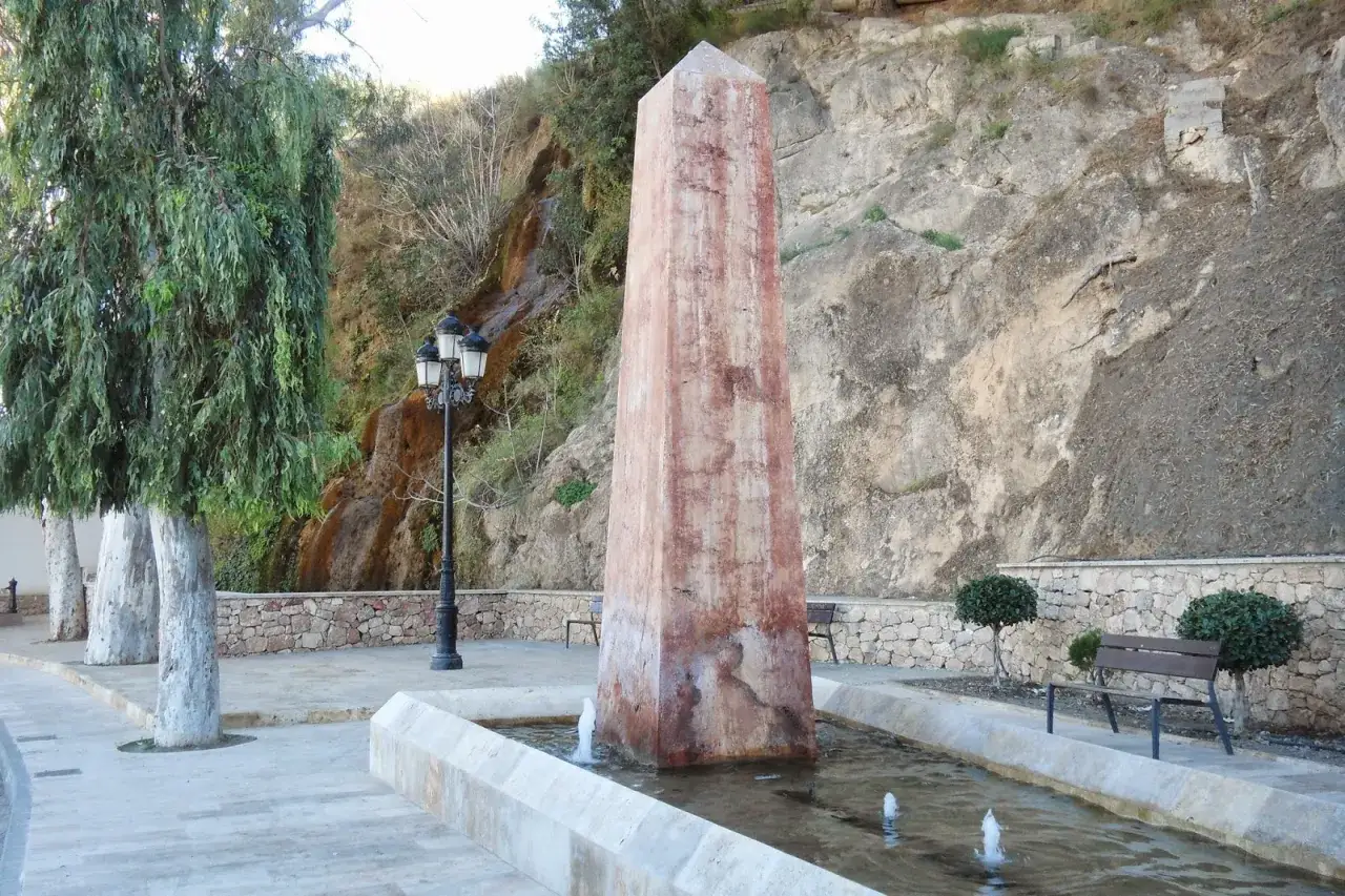 Obelisco en fuente con cascada alhama de almeria al fondo. Árboles y bancos invitan al descanso.