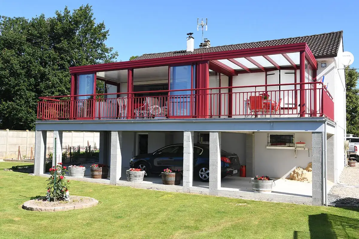 Modèle de véranda sur balcon avec rambarde rouge, offrant un espace de vie lumineux. Une voiture est garée en dessous.