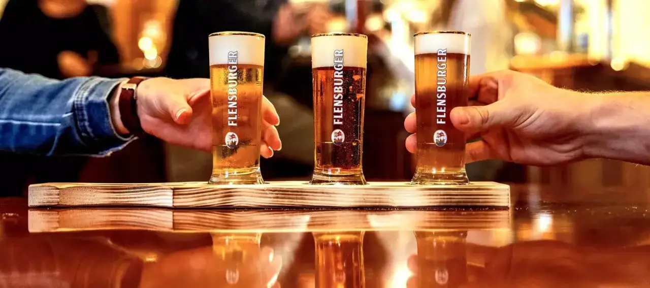 Drei Gl&auml;ser mit goldgelbem Bier und cremigem **Bier schaum** stehen auf einem Holzbrett. H&auml;nde greifen nach den Gl&auml;sern.