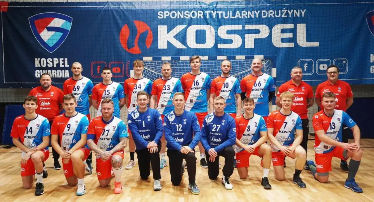 Drużyna Gwardia Koszalin piłka ręczna pozuje do zdjęcia. Zawodnicy w strojach klubowych, z numerami na koszulkach, prezentują się przed banerem sponsora.