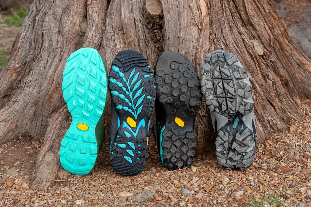 Four shoes lean against a tree, showcasing diverse Vibram sole types. From bright teal to rugged grey, each tread promises grip for any adventure.